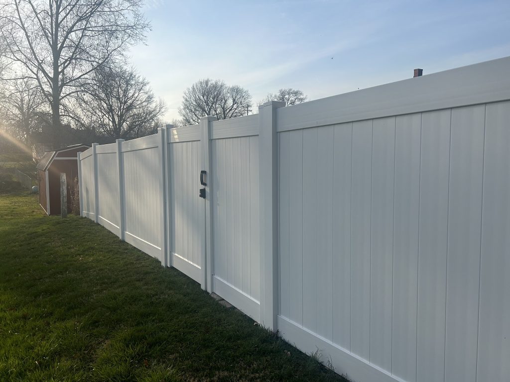 Tall, white vinyl privacy fence close up. There is a gat in the middle of the shot and the sun is setting in the background.